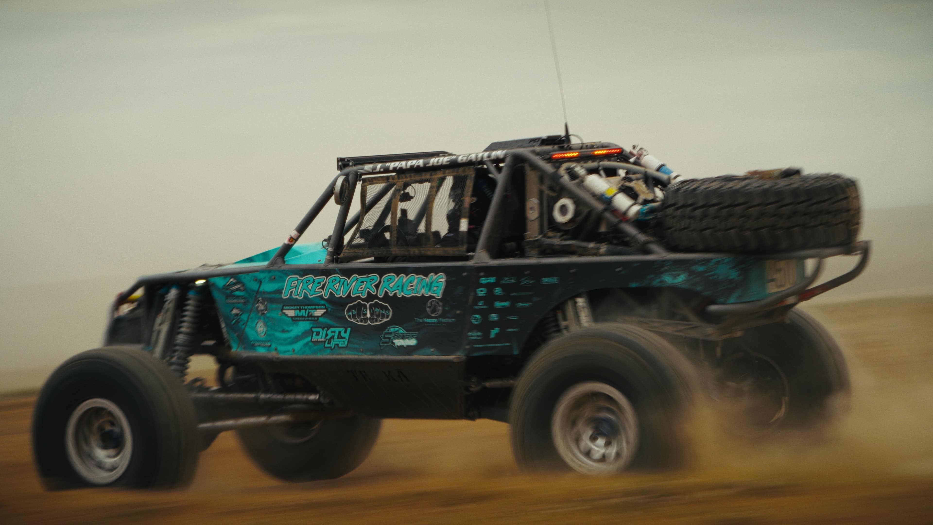 Ground-level chase shot of a race truck airborne over a dirt course.